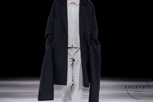 A black dior coat with long to the ground sleeves paired with a zip up collared grey shirt and straight trousers, pink shows with white socks, during Paris Fashion Week Men's Autumn Winter 2024