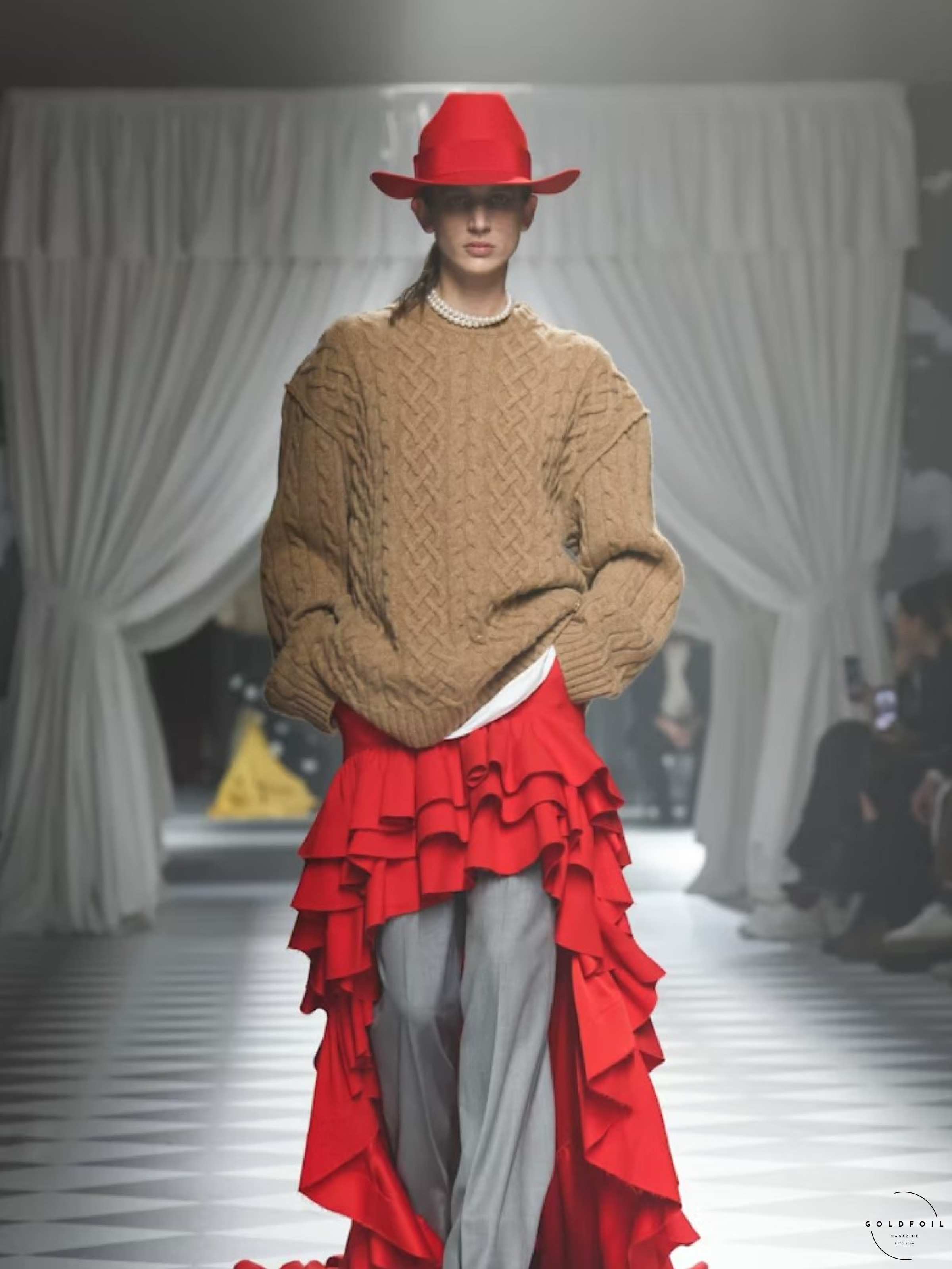 Adrian Appiolaza presented his first runway show for the Moschino brand as part of Milan Fashion Week. Model walking on the runway wearing a long fringe red skirt, with smart flary grey trousers, a white top, pearl necklace and a knitted tan jumper on top with a red hat